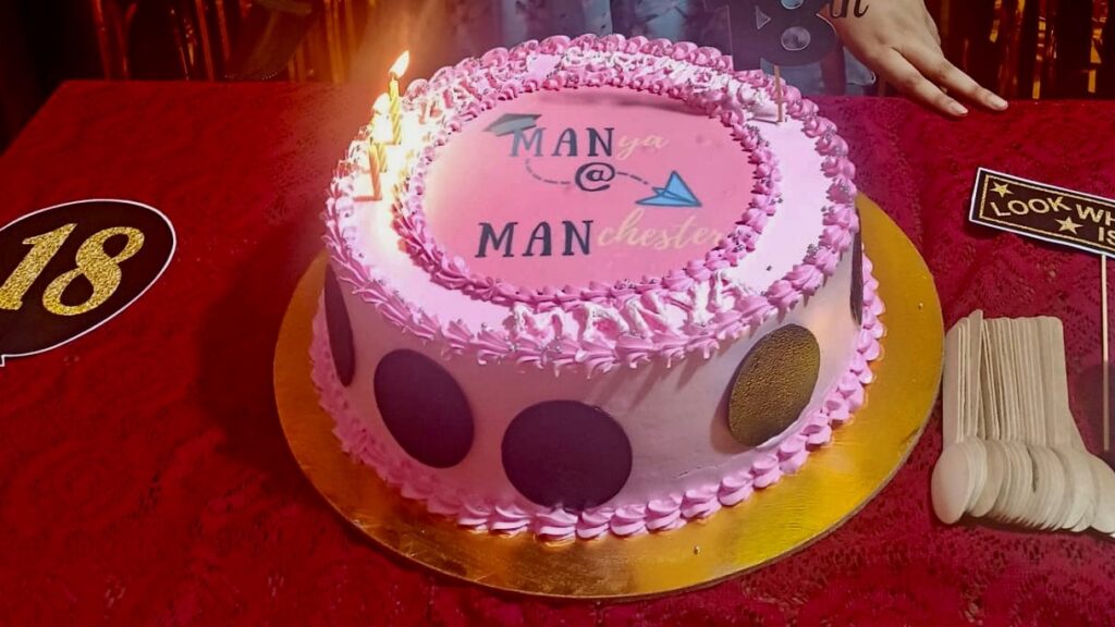 Cake with lighted candles and icing that reads "Manya @ Manchester"