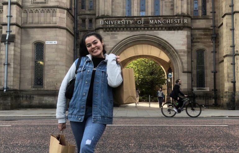 Smiling woman standing in front of building with sign reading "University of Manchester"