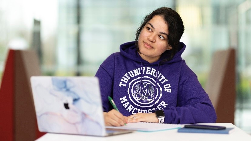 Woman wearing University of Manchester hoodie writing behind an opened laptop