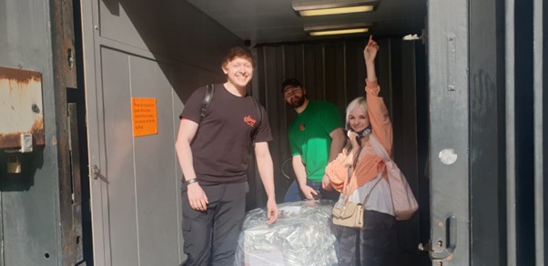 Two male presenting people and a female presenting person stand next to a very large package covered in foil in a grey boxed space.