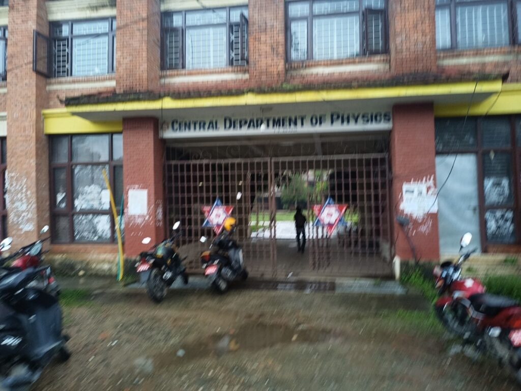 The front of a building with a wide overhang and decorated gates, with the sign 'Central Department of Physics' overhead. There are a number of motorbikes in front of the building.