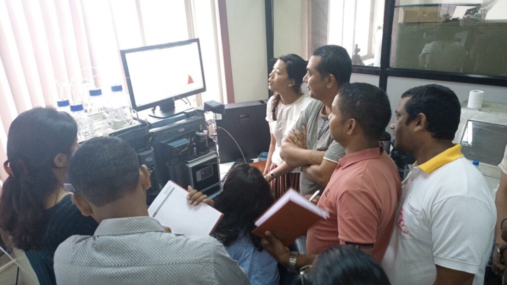 A group of people stand surrounding a computer screen and technical equipment. Some of the people have notebooks to make notes.