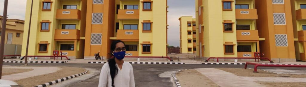 Sanya, wearing a facemask, standing in front of a number of apartment buildings