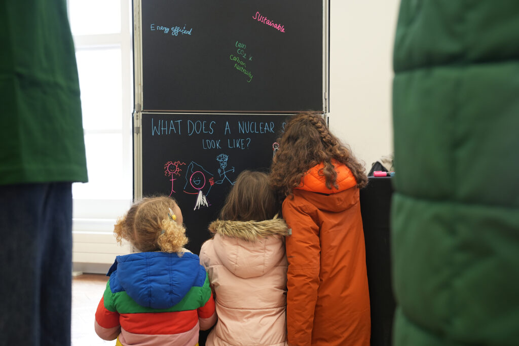 Three children writing on a chalkboard with colourful chalk pens. The chalkboard reads 'What does a nuclear scientist look like?' 