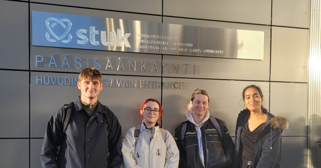 Four people standing outside a building. The sign behind them reads stuk