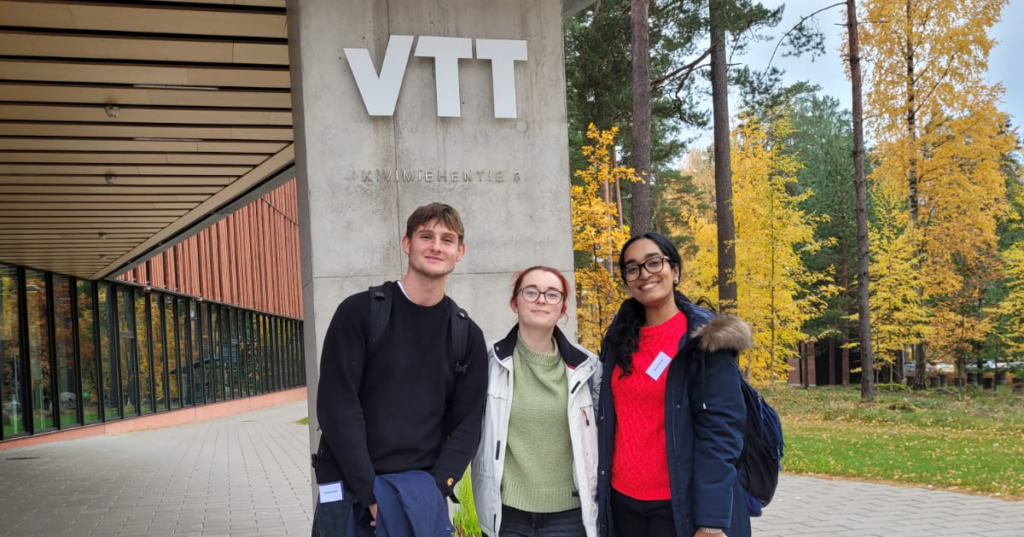 Three people standing outside a building in autumn. The sign behind them reads VTT